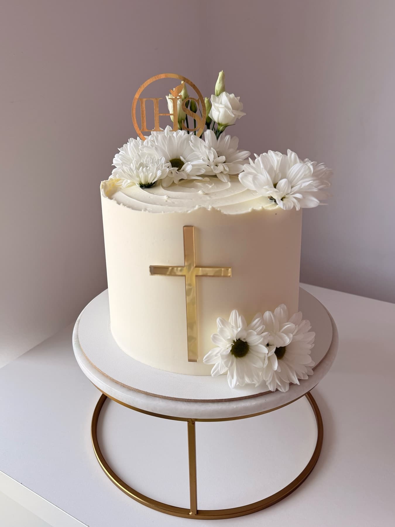 Biały tort komunijny ze złotym krzyżem, monogramem IHS i białymi margerytkami